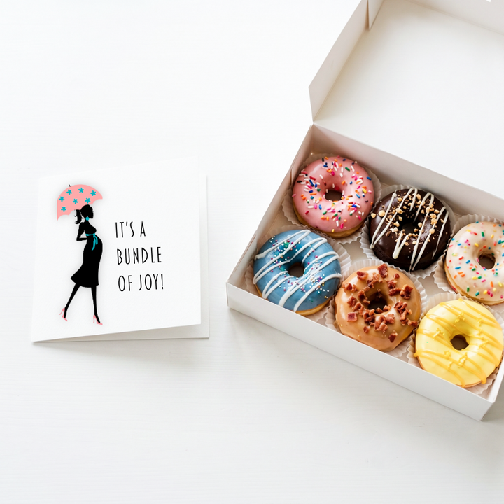 A boxed assortment of six frosted donuts sits beside a white greeting card with a black silhouette of a woman holding a pink polka-dot umbrella and the text, &