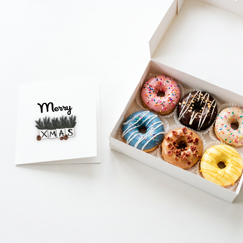 A white gift box containing six assorted frosted donuts sits on a white surface beside a white card that reads 'Merry Xmas' with a small tree illustrations.