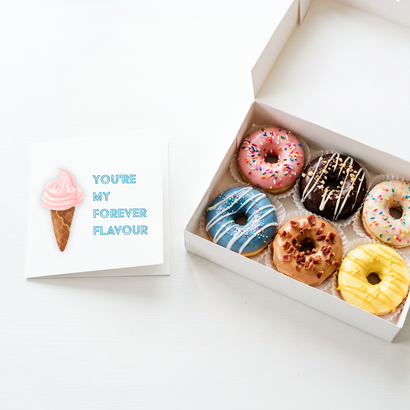 A white gift box containing six assorted donuts with pink, blue, chocolate, and yellow glazes, placed beside a small greeting card that reads 'YOU'RE MY FOREVER FLAVOUR' with a pink ice cream cone illustration.