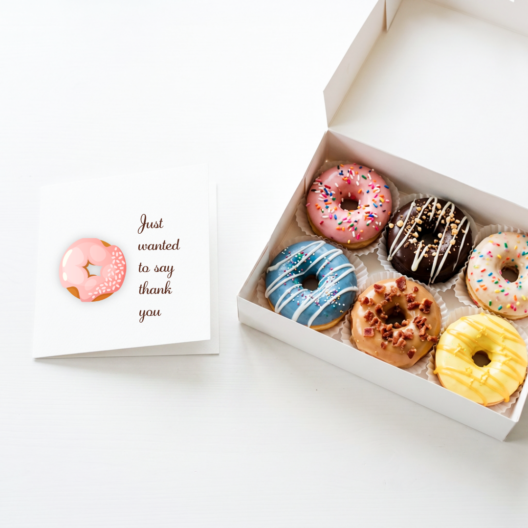 A white greeting card with a pink donut illustration and the text 'Just wanted to say thank you' sits beside an open white box containing six assorted glazed donuts on a white background.
