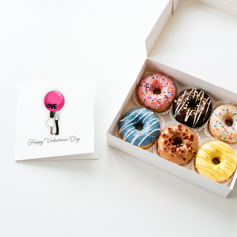 A white gift box containing six assorted frosted donuts next to a square Valentine&