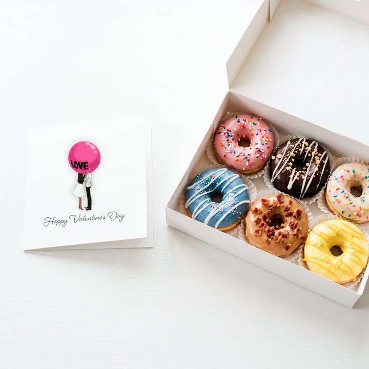 A white gift box containing six assorted frosted donuts next to a square Valentine&